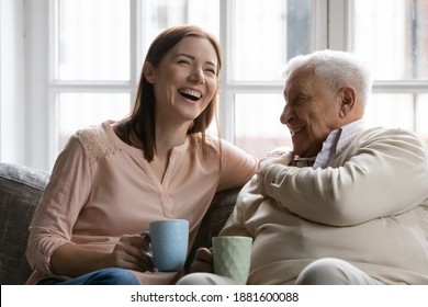 Family members of two generations grown daughter and mature father having fun enjoy tea talk on cozy couch. Attentive young lady caregiver social worker visit support care for glad elderly man patient