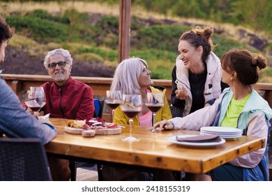 Family members of various ages share laughter and stories over glasses of red wine, enjoying a serene outdoor setting - Powered by Shutterstock