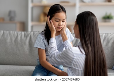 Family misunderstanding concept. Young asian mother calming down her offended daughter after quarrel, sitting on sofa at home, free space. Woman comforting her child