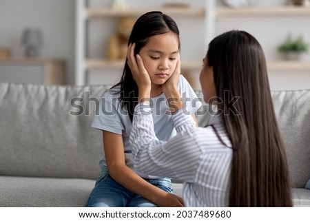 Family misunderstanding concept. Young asian mother calming down her offended daughter after quarrel, sitting on sofa at home, free space. Woman comforting her child