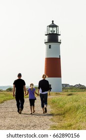 A Family With Mom, Dad And Young Daughter Walking Up A Path Toward A Light House On The East Coast. Concept For Travel In New England And The Atlantic Coast Line.