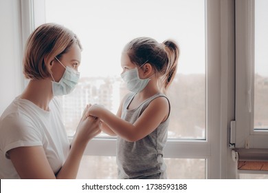 Family Mom And Daughter In Medical Mask. Young Woman And Child Little Girl Sitting By The Window In Protective Masks Against The Virus. Mom Carefully Corrects The Mask On The Face Of The Child      