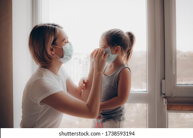 Family Mom And Daughter In Medical Mask. Young Woman And Child Little Girl Sitting By The Window In Protective Masks  Against The Virus. Mom Carefully Corrects The Mask On The Face Of The   Child. 