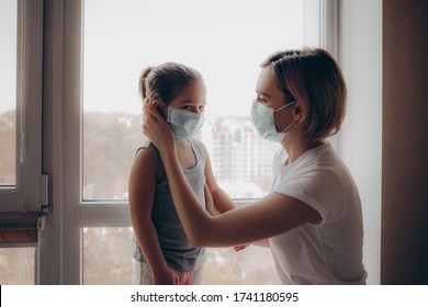 Family Mom And Daughter In Medical Mask. Young Woman And Child Little Girl Sitting By The Window In Protective Masks Against The Virus. Mom Carefully Corrects The Mask On The Face Of The  Child      