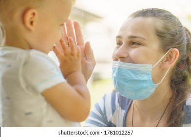 Family Mom And Daughter In Medical Mask.Young Happy Woman And Child Little Boy Sitting By The Window In Protective Masks Against The Virus. Baby Holding Hand On Glass. Mom Strokes The Baby's Face