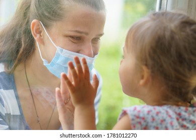 Family Mom And Daughter In Medical Mask.Young Happy Woman And Child Little Boy Sitting By The Window In Protective Masks Against The Virus. Baby Holding Hand On Glass. Mom Strokes The Baby's Face