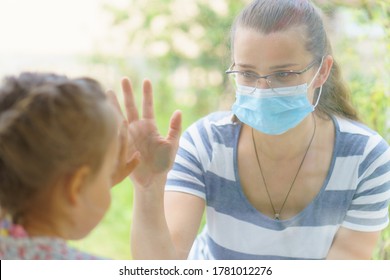 Family Mom And Daughter In Medical Mask.Young Happy Woman And Child Little Boy Sitting By The Window In Protective Masks Against The Virus. Baby Holding Hand On Glass. Mom Strokes The Baby's Face