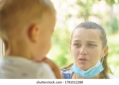 Family Mom And Daughter In Medical Mask.Young Happy Woman And Child Little Boy Sitting By The Window In Protective Masks Against The Virus. Baby Holding Hand On Glass. Mom Strokes The Baby's Face