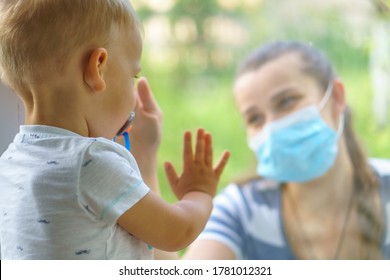 Family Mom And Daughter In Medical Mask.Young Happy Woman And Child Little Boy Sitting By The Window In Protective Masks Against The Virus. Baby Holding Hand On Glass. Mom Strokes The Baby's Face