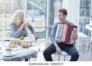 Family moments at home.Young handsome male playing accordion for beautiful mother. Attractive mature Caucasian woman looking at adorable son with love drinking wine, lunch time at stylish luxury home. - Powered by Shutterstock