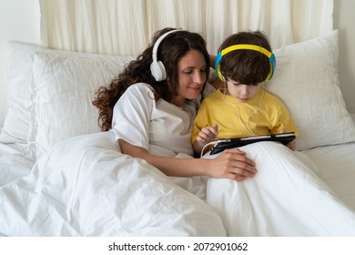 Family Morning In Bedroom: Mom Look At Little Kid Son Play Games On Tablet Computer Or Watch Video Cartoons Wearing Headphones And Listening To Music Lying With Child In Bed Under Blanket On Weekend