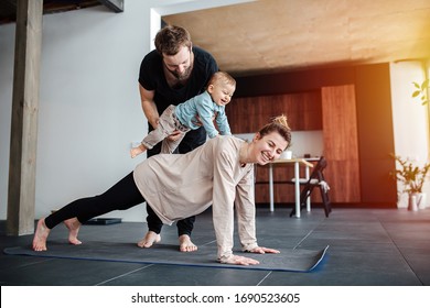 Family Morning Exercise. Mother Doing Plank, Father Holding Their Baby On Her Back, So He Would Ride Her, Including Child In Activity. Family Quarantine, Domestic Life In Self-isolation. Sunset Light