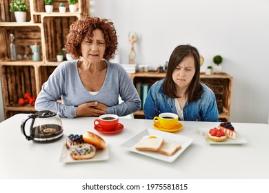 Family Of Mother And Down Syndrome Daughter Sitting At Home Eating Breakfast With Hand On Stomach Because Indigestion, Painful Illness Feeling Unwell. Ache Concept. 