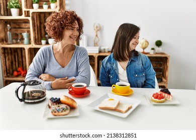 Family Of Mother And Down Syndrome Daughter Sitting At Home Eating Breakfast Looking Away To Side With Smile On Face, Natural Expression. Laughing Confident. 