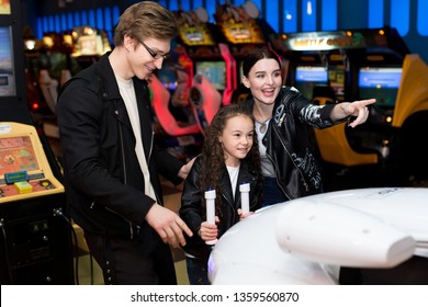 Family Mother, Father, Daughter Play Arcade Games On The Computer Machines At An Amusement Park. Children And Adults Play On The Slot Machines, Attractions In The Shopping Center.