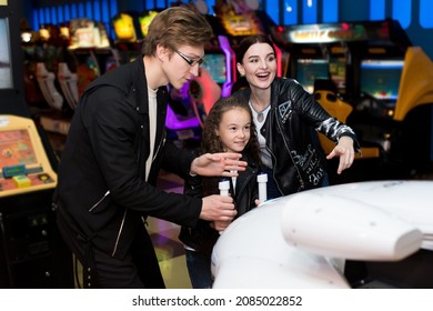 Family Mother, Father, Daughter Play Arcade Games On The Computer Machines At An Amusement Park. Children And Adults Play On The Slot Machines, Attractions In The Shopping Center.