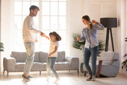 Family Mother Father And Preschool Adorable Daughter In Living Room Moving Dancing To Music Little Girl Holding Father Hand Having Fun Enjoy Time With Parents At Home. Funny Leisure Activities Concept