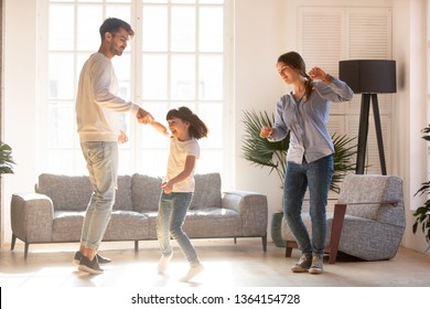 Family Mother Father And Preschool Adorable Daughter In Living Room Moving Dancing To Music Little Girl Holding Father Hand Having Fun Enjoy Time With Parents At Home. Funny Leisure Activities Concept