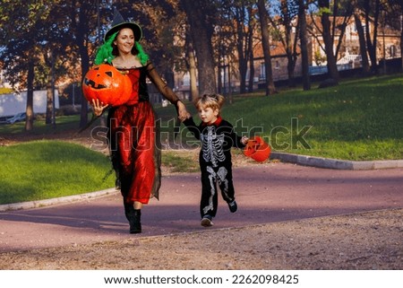 Family with mother and little boy run in Halloween disguise costumes of skeleton, witch hold bucket for candies over town street