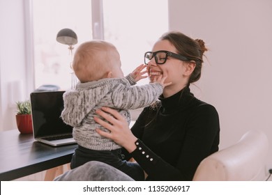 Family, Mother Working With Child. Young Mother Working At Home Sitting In Chair Holding Her Little Son And Smiling To Him. Mom Is Using A Computer And Spending Time With Her Cute Baby Boy At Home