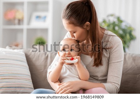 family, motherhood and people concept - happy smiling mother kissing little baby playing with teething toy or rattle at home