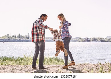 Family Is Not An Important Thing. It's Everything. Parents Standing With Their Kid, Holding His Hands And Lifting Him Up. Summer Landscape. Back View