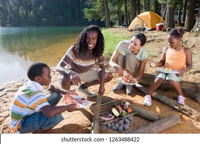 Family On Camping Trip By Lake Cooking Food On Barbecue