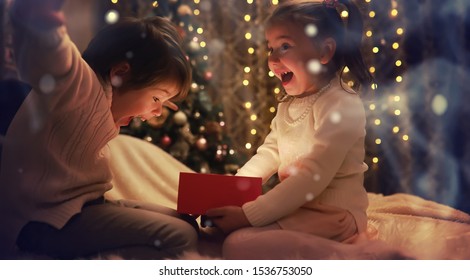 Family On Christmas Eve At Fireplace. Kids Opening Xmas Presents. Children Under Christmas Tree With Gift Boxes. Decorated Living Room With Traditional Fire Place. Cozy Warm Winter Evening Home.