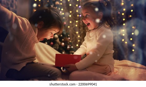 Family On Christmas Eve At Fireplace. Kids Opening Xmas Presents. Children Under Christmas Tree With Gift Boxes. Decorated Living Room With Traditional Fire Place. Cozy Warm Winter Evening Home.