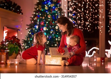 Family On Christmas Eve At Fireplace. Mother And Little Kids Opening Xmas Presents. Children With Gift Boxes. Living Room With Traditional Fire Place And Decorated Tree. Cozy Winter Evening At Home.