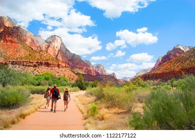 Family On Hiking Trip In The Mountains Walking On Pathway. People Wearing Backpacks Hiking In Red Mountains Zion National Park, Utah, USA