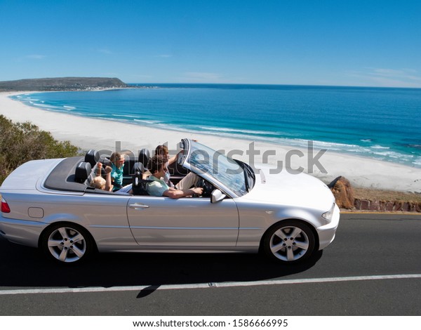 Family on holiday driving convertible on road\
with beach and ocean in\
background