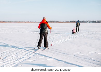 Family On Ski Trip. Young Woman, Man And Child Are Skiing In Winter On Frozen River Near Forest, Man Is Sledding Child. 