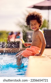 Family On Summer Holiday With Boy Eating Ice Lolly Splashing At Edge Of Swimming Pool