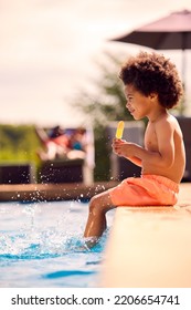 Family On Summer Holiday With Boy Eating Ice Lolly Splashing At Edge Of Swimming Pool