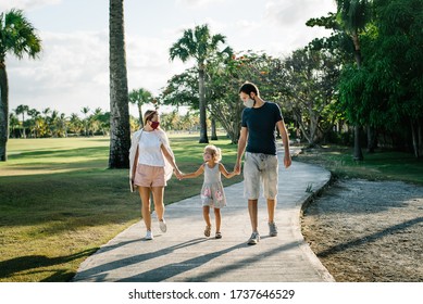 Family On A Walk In The Park In Protective Masks Against The Corona Of The Virus Covid-19. Sunny Day On The Background Of Palm Trees.
