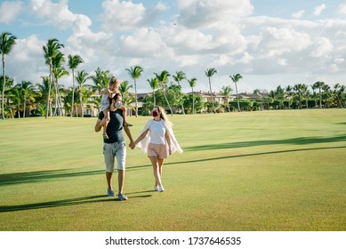 Family On A Walk In The Park In Protective Masks Against The Corona Of The Virus Covid-19. Sunny Day On The Background Of Palm Trees.
