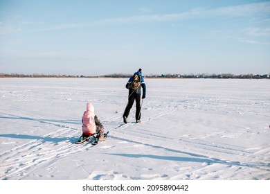 Family on winter walk. Young man and child are skiing in winter on frozen river near forest, man is sledding child in snow. 