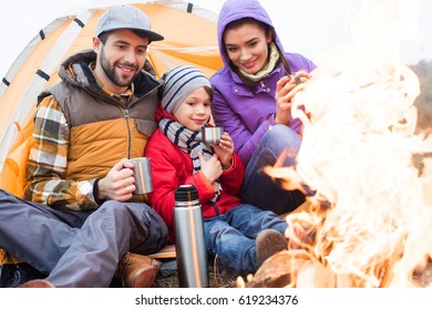 Family with one child sitting near tent and drinking hot tea from thermos while resting near burning fire in countryside - Shutterstock ID 619234376