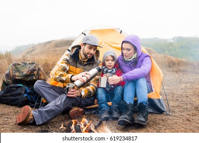 Family with one child sitting near tent and drinking hot tea from thermos while resting near burning fire in countryside - Shutterstock ID 619234817
