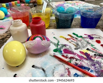 A Family Painting Easter Eggs With Crayola Paint. Dyed Eggs With A Rainbow Painted On, Colorful Paint Streaks, Dye Cups, Paint Brushes, And Paper Towels. 