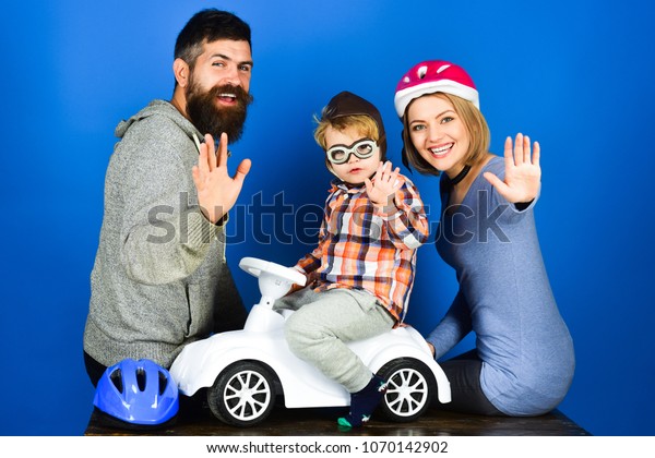 Family, parenthood, adoption and people concept -\
happy mother, father with protective helmet, son ride on toy car\
waving hands. Boy in helmet or hat and glasses driving race car.\
Vacation and leisure