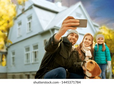 Family, Pets And People Concept - Happy Mother, Father And Little Daughter With Beagle Dog Taking Selfie By Smartphone Outdoors Over House In Autumn Background