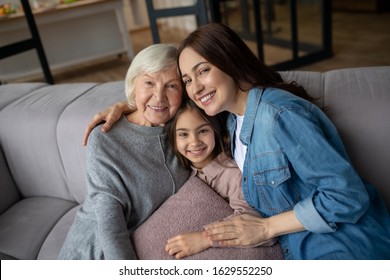 Family Photo. Girl, Mother, Grandmother Sitting Embraced On The Couch, Looking Straight Ahead, Smiling And Happy.