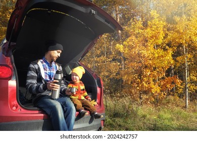 Family Picnic Outdoor, Road Trip In Autumn Season. Father And His Little Child Sitting Inside Car Trunk Near Rural Road In Autumn Forest  And Drinking Tea From Thermos.