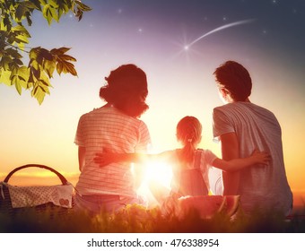 Family Picnicking Together. Young Mother, Father And Child Daughter Girl Sitting Together On Green Grass In Summer Park And Making A Wish By Seeing A Shooting Star.