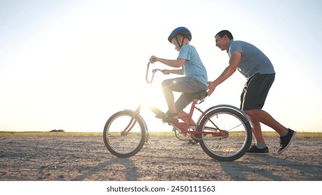 family play in the park. father teaching son to ride a bike. happy family kid dream concept. son learn to ride bike silhouette. father supporting child son riding bike summer sunset in the park - Powered by Shutterstock