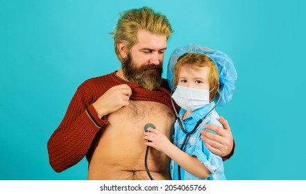 Family Playing In Doctor And Patient. Son In Medical Mask With Stethoscope Listening To Heartbeat Of His Father.