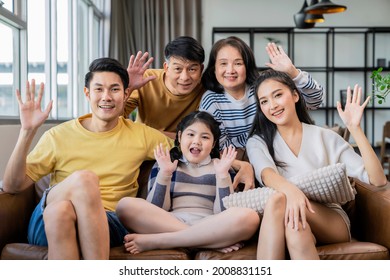 Family Portrait Asian Multi Gneration  Parent Grandparent And Grandchild Sit Relax Together Smile Hand Wave To Say Hi Look At Camera On Sofa At Living Room Home Interior Background