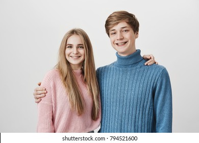 Family portrait of brother and sister against gray background. Young hipster fair-haired man dressed in blue sweater embracing smiling beautiful blonde sister with long hair. Smiling positive couple